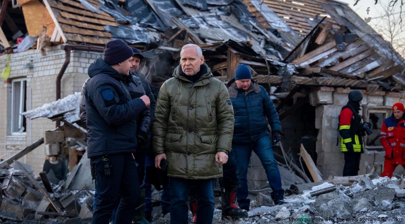 A medium shot shows Ihor Terekhov, the Mayor of Kharkiv, wearing an olive-green puffer jacket and standing amidst heavy debris. He has a concerned expression while looking toward someone off-camera. In the background, a residential house is severely damaged, with its roof partially collapsed and structural beams exposed. Several men in winter clothing, a rescuer in a high-visibility uniform, and a medical worker are also present at the scene. A photo credit to "Ukrinform" is in the bottom-right corner.