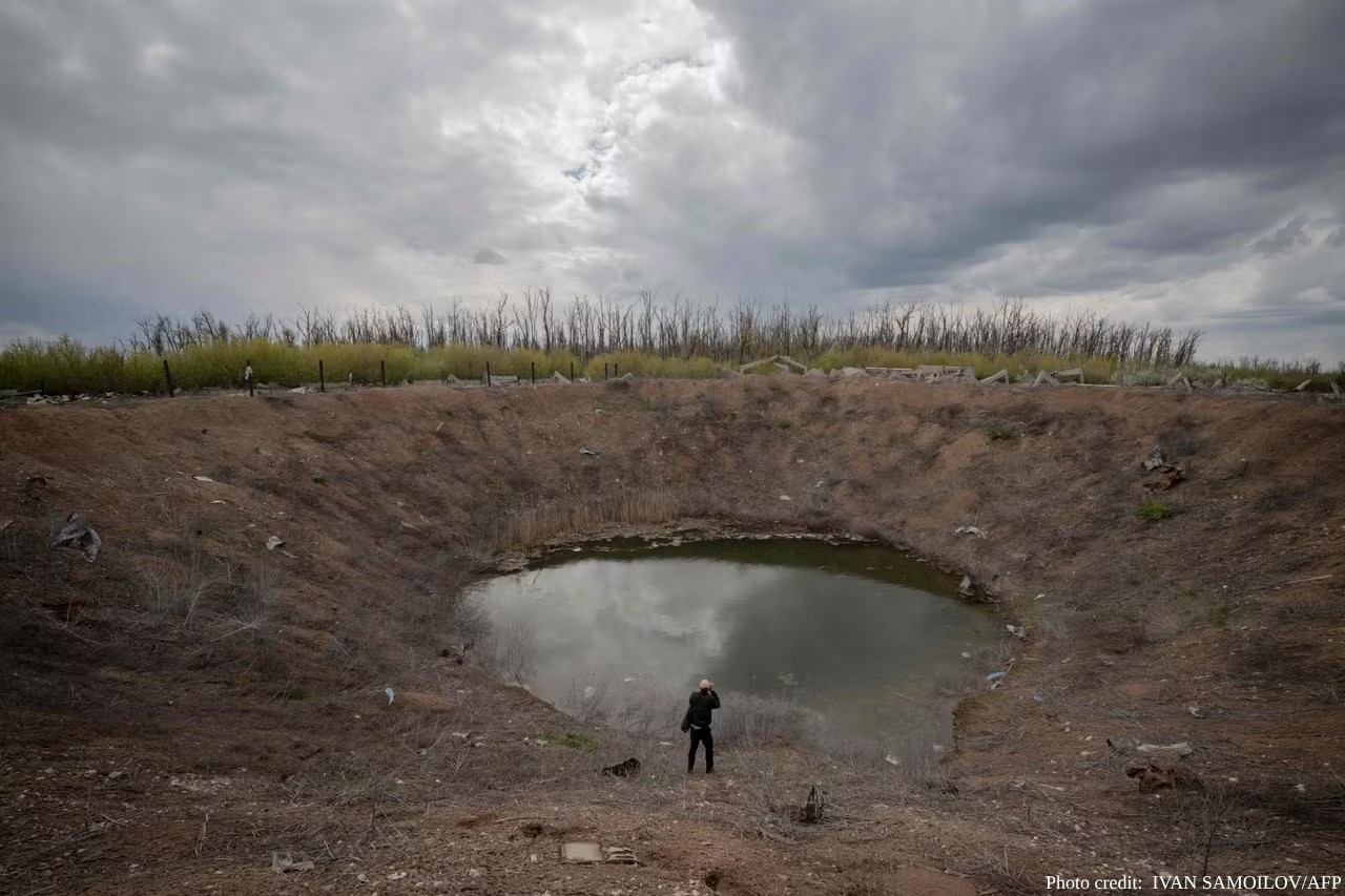 A person stands at the bottom of a massive, bowl-shaped crater in the Kharkiv region, which has partially filled with stagnant water reflecting the overcast sky. The steep, earthen walls of the crater are sparsely covered with dry brush. In the background, a line of thin, leafless trees stands against a heavy, grey cloud cover. The photo, taken on May 1, 2026, is credited to IVAN SAMOILOV/AFP in the bottom-right corner.