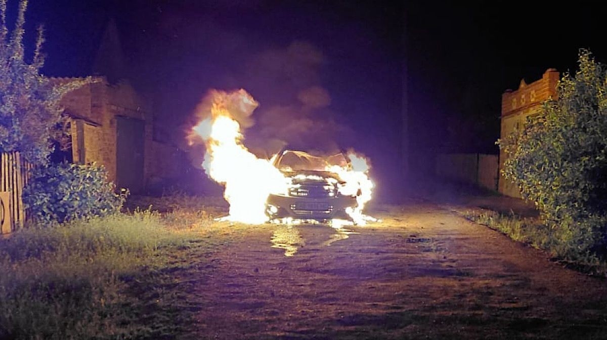 A nighttime photo of a passenger car completely engulfed in bright, roaring flames on a dark dirt road. The fire is most intense at the front and side of the vehicle, casting a strong light on the surrounding ground and nearby buildings. Thick smoke rises into the night sky. The National Police of Ukraine logo is positioned in the bottom-right corner.