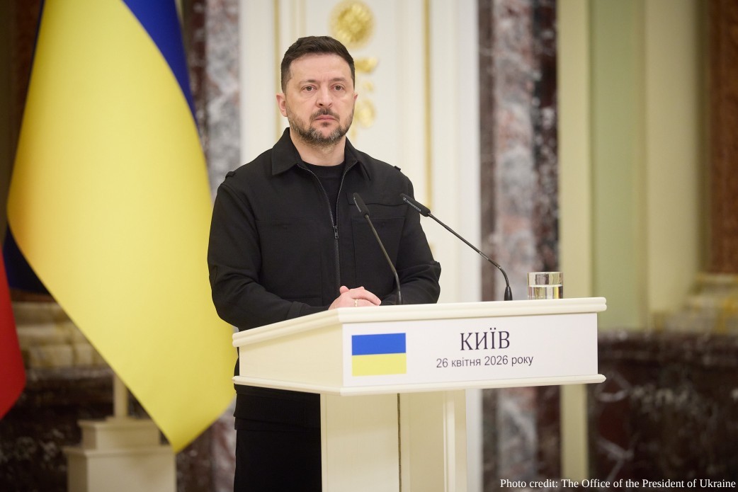 A medium shot of President Volodymyr Zelenskyy standing behind a white podium in a formal hall. He is wearing a black zip-up jacket and has a serious expression. The podium features the Ukrainian flag and the text "КИЇВ, 26 квітня 2026 року" (Kyiv, April 26, 2026). A large Ukrainian flag is visible in the background, and a photo credit to "The Office of the President of Ukraine" is in the bottom-right corner.