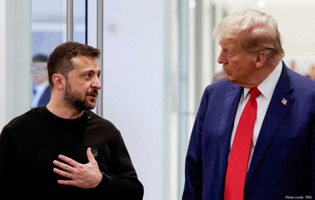 A photograph showing Ukrainian President Volodymyr Zelenskyy on the left and Donald Trump on the right. Zelenskyy is wearing a black long-sleeve shirt with a small Ukrainian trident emblem, with his hand on his chest, appearing to speak. Trump, wearing a dark blue suit, white shirt, and red tie with an American flag pin, is looking at Zelenskyy. The background is a bright, blurred indoor space.