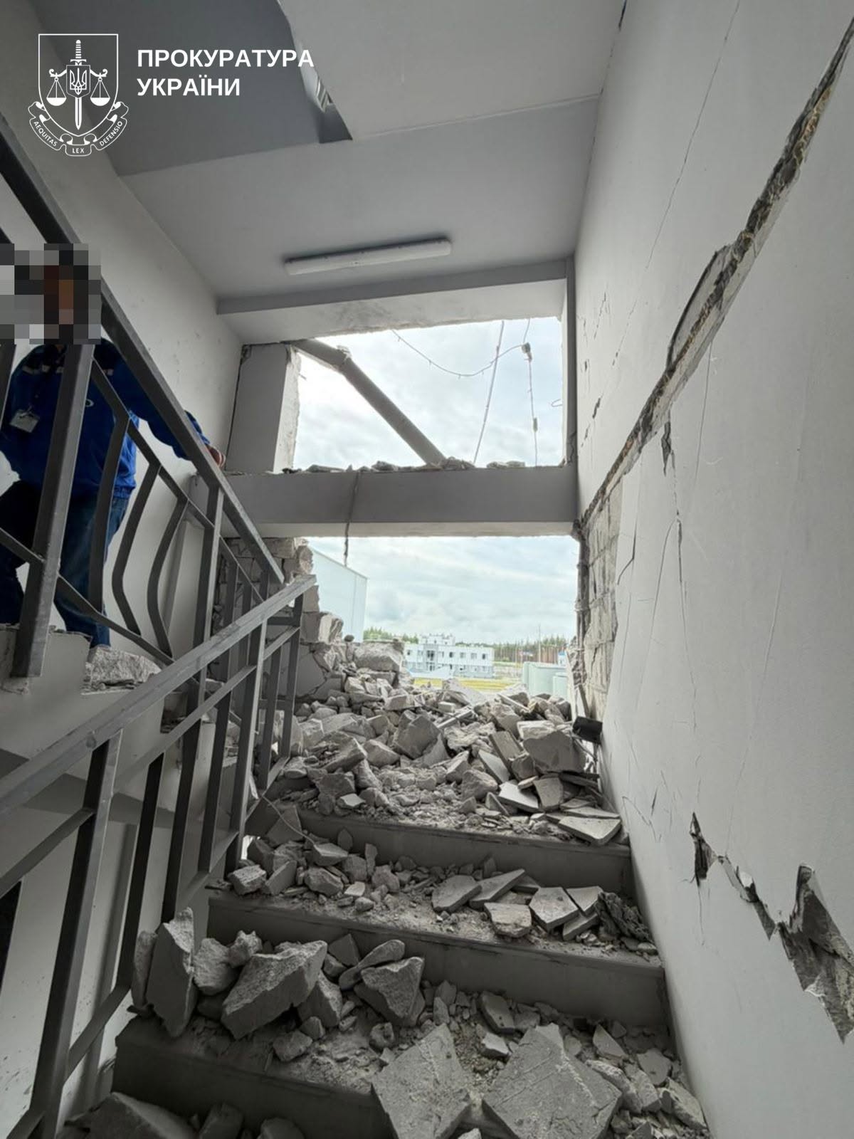 A stairwell inside a building in Ukraine is covered in rubble and concrete debris following an attack that blew out an exterior wall and cracked the adjacent structure. A person with a blurred face stands on the stairs, and the emblem of the Prosecutor General's Office of Ukraine is visible in the upper-left corner.