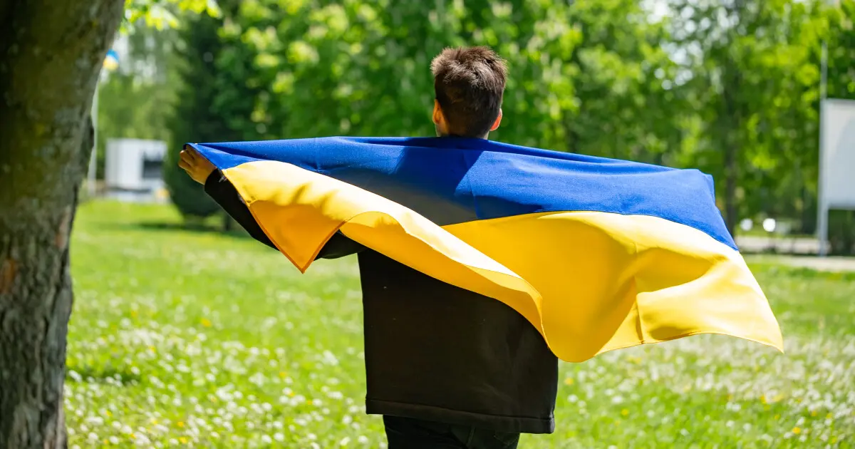 Юнак з прапором України на плечах стоїть на фоні зеленої галявини.