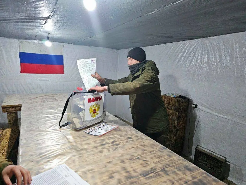 Чоловік кидає бюлетень до виборчої скриньки на дільниці в тимчасово окупованому місті; на стіні – прапор Росії.