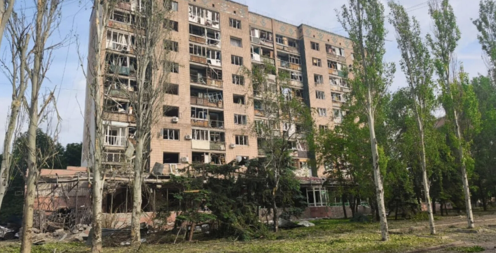 Пошкоджена внаслідок російського обстрілу багатоповерхівка в Краматорську. На зображенні видно посічені дерева, вибиті вікна та зруйнована конструкція.